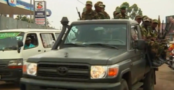 Military personnel ride in a green pickup truck on a busy street, surrounded by traffic, including a white van in the background.