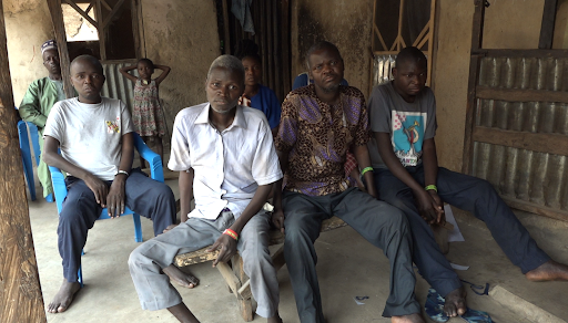 A group of six people sitting and standing in a small room with a bare, rustic interior.