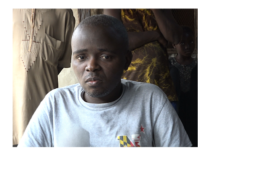 A person in a gray shirt sits indoors, with others standing in the background.