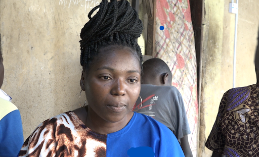 A woman in a blue and patterned top stands in front of a wall and a person facing away, with patterned fabric in the background.