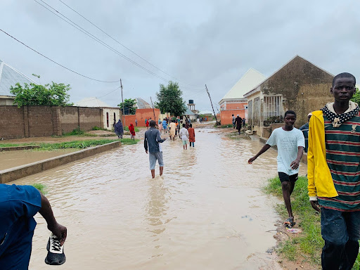 People walking through a flooded street in a residential area after heavy rain. Overcast sky above.