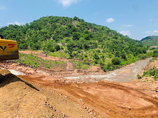 A construction site with a muddy river flowing through a hilly, green landscape under a blue sky.