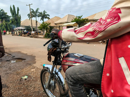 Person riding a motorcycle on a street lined with houses and palm trees under a cloudy sky.