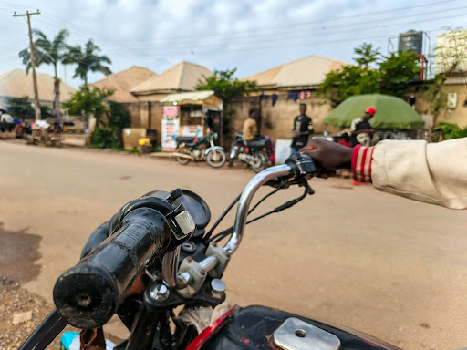 View from a motorcycle handle, with a person riding on a quiet street lined with small shops and greenery in the background.