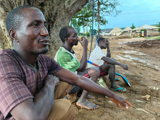 Three men sit under a tree in a rural setting, looking contemplative. Huts are visible in the background.