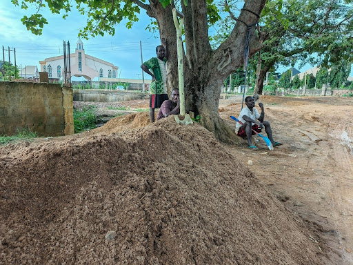 Three people resting by a tree near a dirt pile, with a church visible in the background.