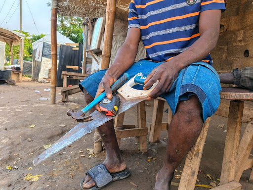 Person sitting on a wooden bench holding a saw and hammer, wearing a striped shirt and denim shorts in an outdoor setting.