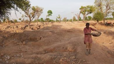 A person in a dusty landscape carries a large bowl. Dirt and sparse trees surround them under a clear sky.