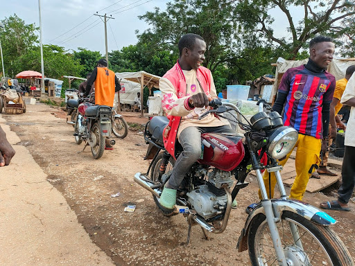 Man on a motorcycle talking with friends on a dirt road, with market stalls and trees in the background.