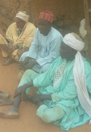 Three men in traditional attire sit on the ground outside, engaging in conversation.