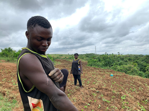 Two people standing in a field, one adjusting their shirt, with cloudy skies overhead.