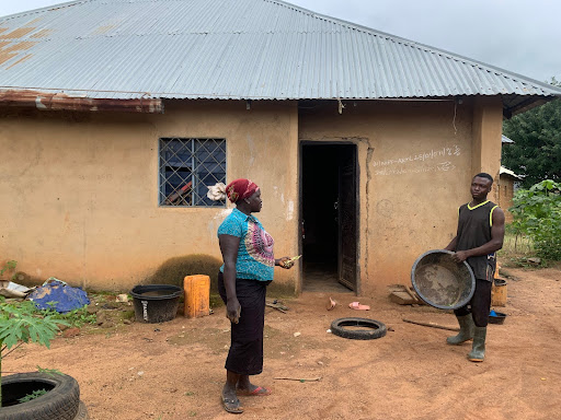 Two people stand outside a small building. One holds a large pan while the other gestures, surrounded by scattered items and tires.