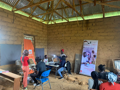 People in a rustic classroom setting with laptops, engaged in instructional activities. Banners with text and images are displayed.