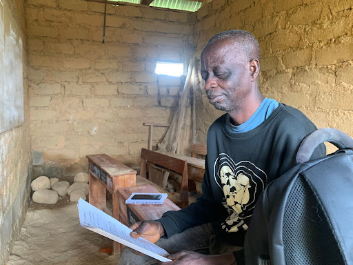 Man sitting in a rustic room, reading a paper with a phone nearby. He's wearing a black sweater, sitting on a bench beside a backpack.
