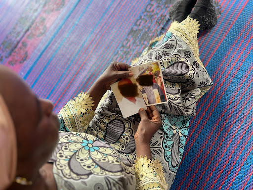 Person in patterned clothing sits on a mat, holding a small photo with two people in it, faces blurred.