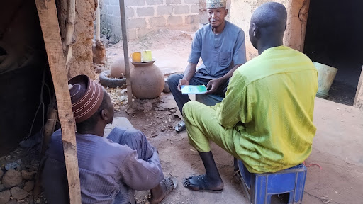 Three men sit and talk outside in a rustic setting, one holding a booklet.