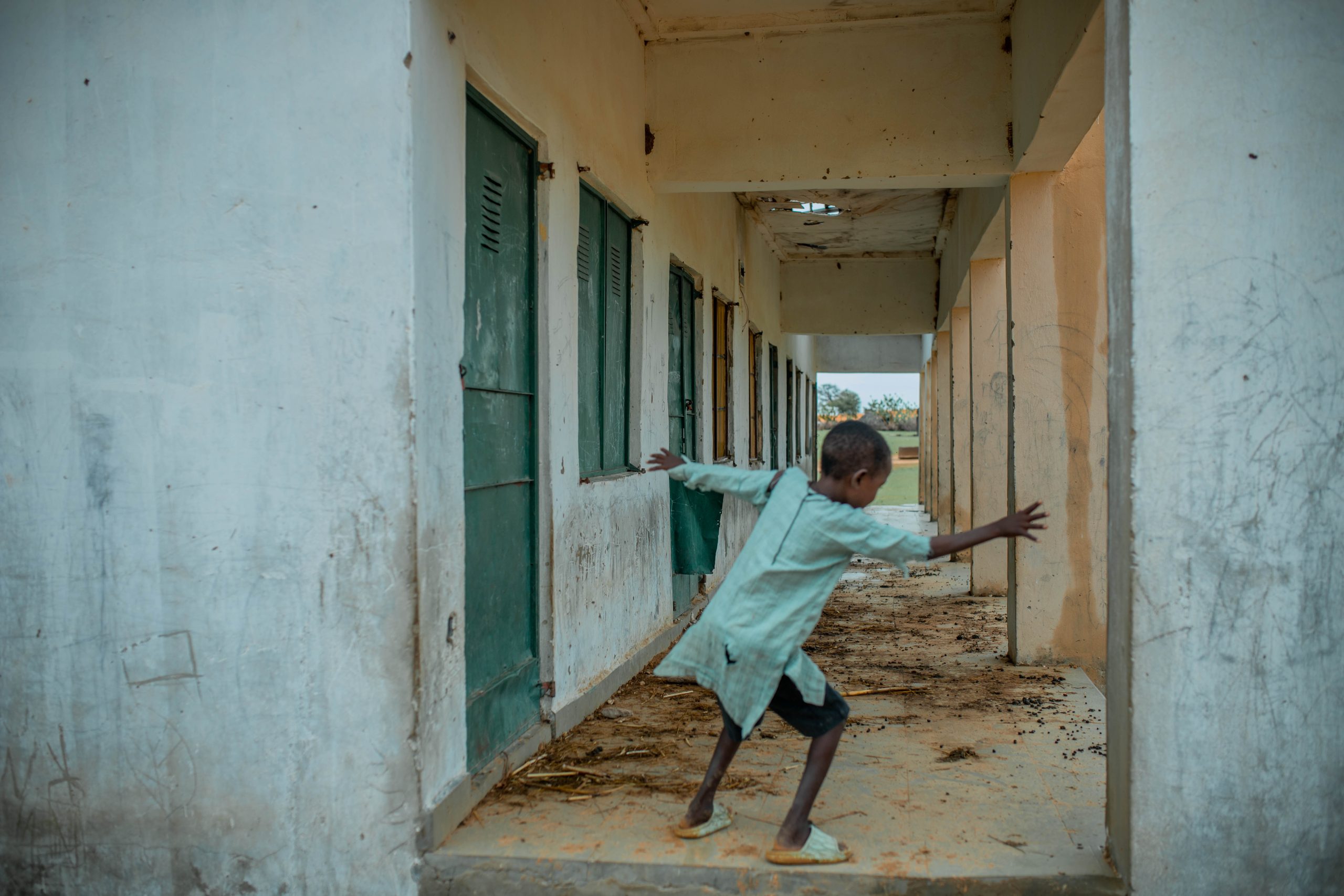 Child in a green shirt playfully leans against a worn corridor wall with green doors, surrounded by scattered debris.