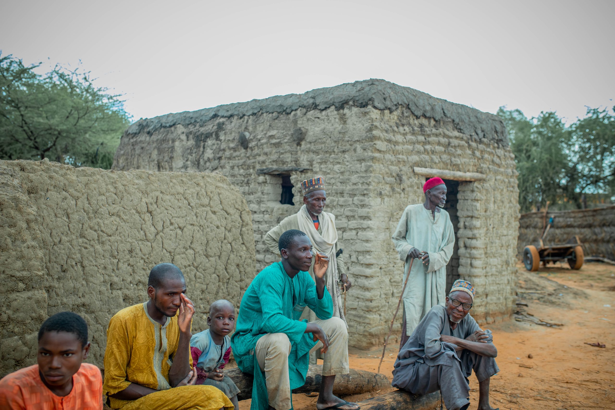 A group of people sitting and standing by mud huts in a rural setting, with trees in the background.