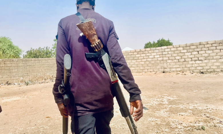 boko haram attack in lake chad Back view of a person walking outdoors with a shotgun slung over their back and holding another in hand, near a brick wall.