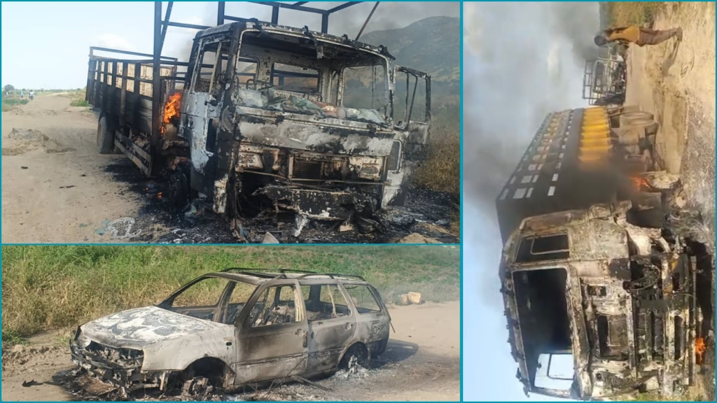 Burned vehicles on a dirt road, showing extensive fire damage to a truck and car, with smoke and some flames visible.