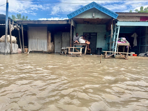 Flooded street with people sitting outside a building, surrounded by water.