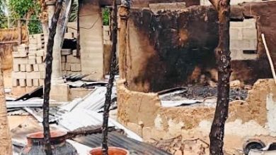 Charred remains of a burnt house with damaged walls and roof. Clay pots sit in front of the rubble, among scorched beams and debris.