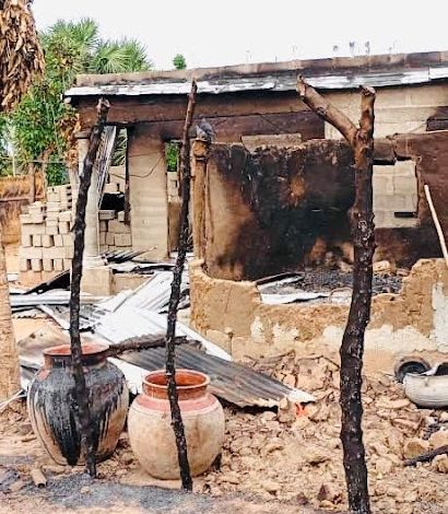 Charred remains of a burnt house with damaged walls and roof. Clay pots sit in front of the rubble, among scorched beams and debris.