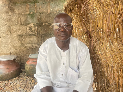 Man in white attire, wearing glasses, sitting on gravel with pots and a straw wall background.