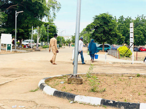 People walking on a sunny day near trees and parked cars, one carrying a blue bucket.