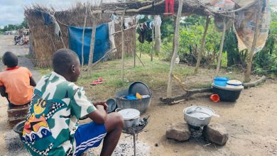 Two children sitting outside a makeshift shelter with cooking pots on a stone fire, surrounded by greenery and laundry hanging above.