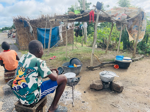 Two children sitting outside a makeshift shelter with cooking pots on a stone fire, surrounded by greenery and laundry hanging above.