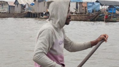 Person in a hoodie paddling a boat on a river, with stilt houses in the background.