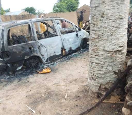 Burned-out van on a dirt road, surrounded by debris and a tree in the foreground.
