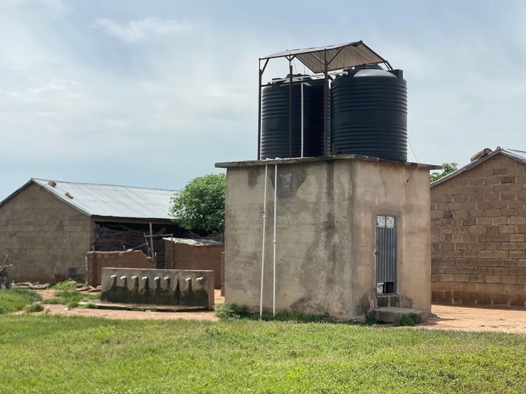 Concrete structure with two large black water tanks on top, adjacent to brick buildings on a grassy area under a cloudy sky.