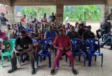 A group of people seated and standing under a shelter, attentively facing forward in an outdoor community gathering.