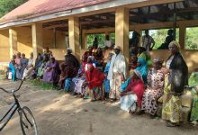 A group of people, mostly women, sit and stand outside a building with a rusty roof. A bicycle is parked in the foreground.