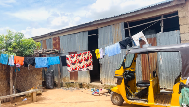 Yellow tuk-tuk parked outside a corrugated metal house, clothes hanging on a line. Sunny day with blue sky.