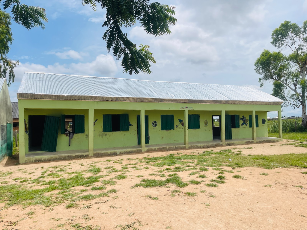 A small, single-story green school building with open doors and windows under a cloudy sky, surrounded by trees and grass.