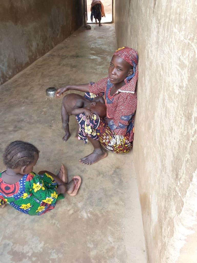 A woman sitting in a corridor holding a baby, with another child nearby. A person is in the background.