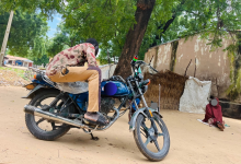 Person on a motorcycle beside a tree, with another person sitting on the ground nearby, against a backdrop of a rustic building and huts.