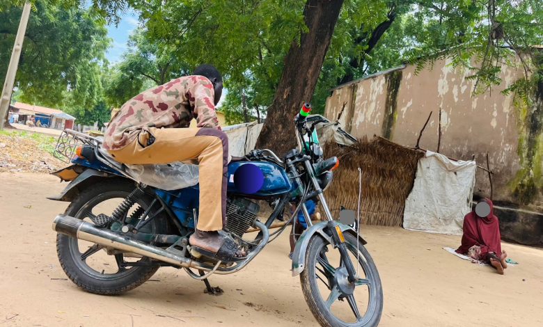 Person on a motorcycle beside a tree, with another person sitting on the ground nearby, against a backdrop of a rustic building and huts.