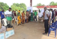 A group of people standing in a semicircle outdoors, some bowing their heads. Boxes and chairs are near them.