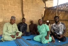 A group of men sitting on a blue mat inside a rustic shelter with straw walls, engaged in conversation.