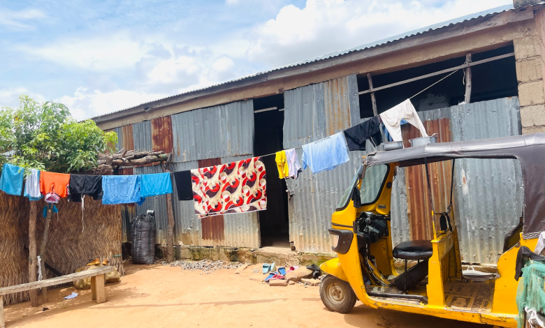 Yellow tuk-tuk parked outside a corrugated metal house, clothes hanging on a line. Sunny day with blue sky.
