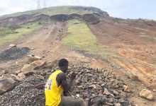 Person in a yellow jersey sitting on rocks with a hill and telecommunications towers in the background under a cloudy sky.
