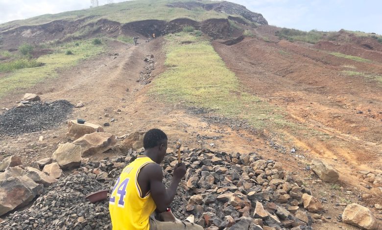 Person in a yellow jersey sitting on rocks with a hill and telecommunications towers in the background under a cloudy sky.