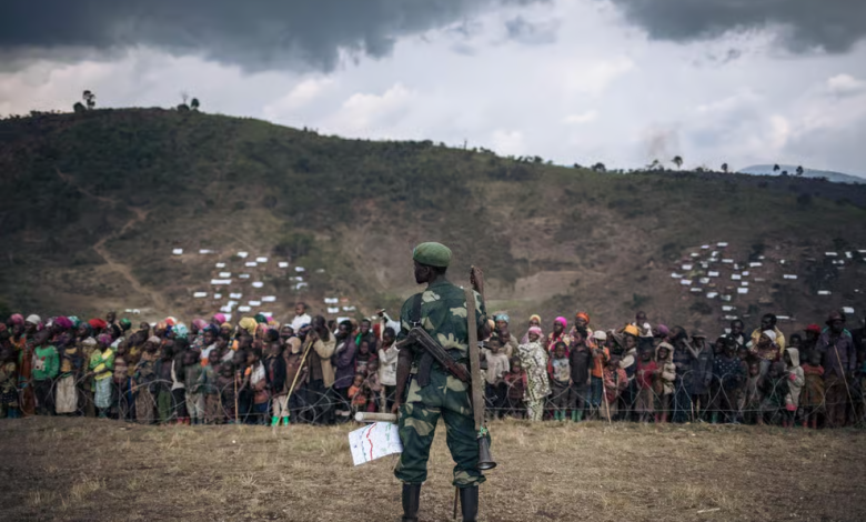Soldier stands in front of a large crowd behind a fence, with hills and tents in the background under a cloudy sky.