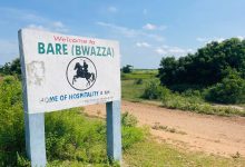 Sign for Bare (Bwazza), "Home of Hospitality," amid greenery under a blue sky. Dirt path leading into the distance.