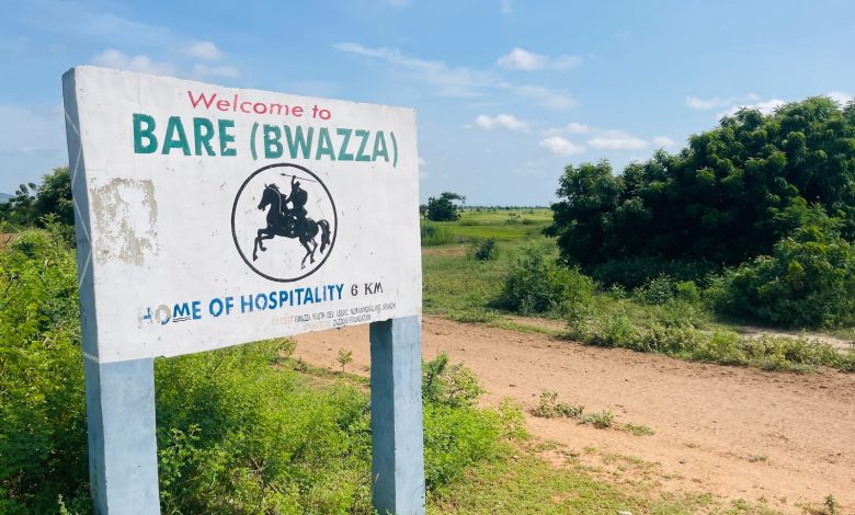 Sign for Bare (Bwazza), "Home of Hospitality," amid greenery under a blue sky. Dirt path leading into the distance.