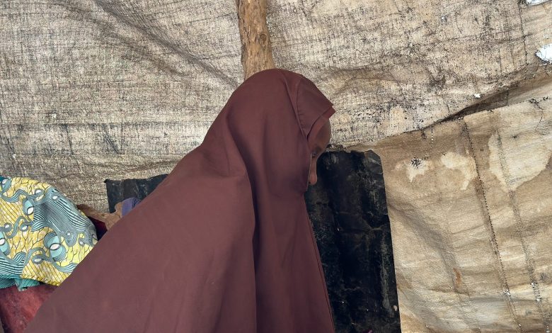 Person in a brown cloak sitting inside a rustic, tarp-covered shelter.